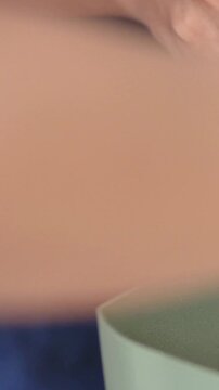 Vertical video: Tilting pink scoop, hands adding soil into matte green-gray planter, blue fabric