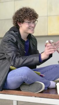 Vertical video: Glancing man in jacket sipping paper cup, scrolling red phone on bench laughing