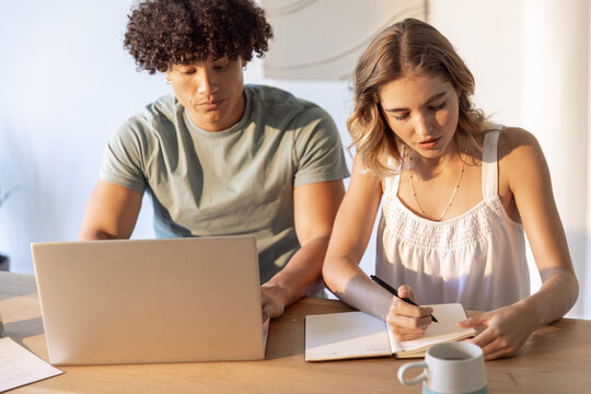 Male and female study partners sitting at wooden table using silver laptop and writing notes