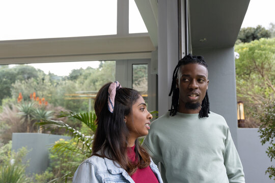 Diverse couple standing and talking at modern doorway with glass panels, succulents, denim jacket