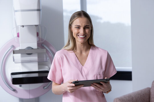 Adult woman in pink scrubs standing in imaging room beside mammography unit holding tablet smiling