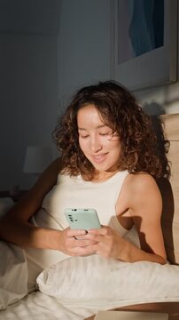Vertical shot of young adult Asian woman lying in bed, scrolling on smartphone and smiling then looking away in morning sunlight. Footage is perfect for social media marketing or wellness campaigns