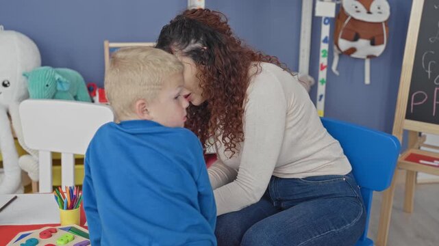 Woman leans close and whispers to a little boy over crayons and wooden shape toys on a small table in a preschool classroom; calm instruction.