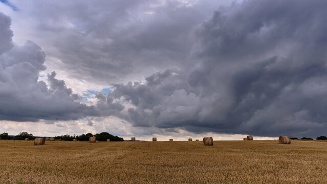 View of round hay bales scattered across a harvested wheat field under dramatic dark storm clouds Skane County, Sweden.