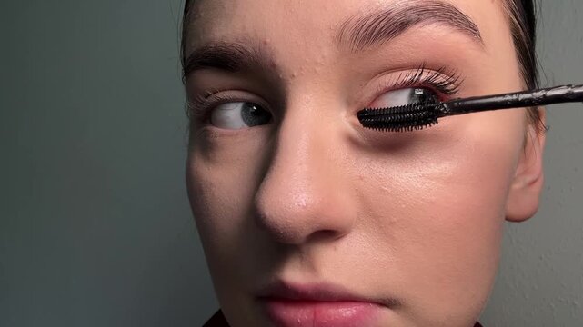 Young woman applying mascara onto her natural eyelashes, demonstrating precise eye makeup enhancement, showcasing daily beauty ritual, facial cosmetic preparation, personal grooming process.