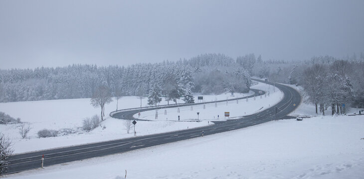 View of a winding road through the snow-covered Eifel landscape with frost-laden pine trees and white fields under an overcast winter sky in Kelberg, Rhineland-Palatinate, Germany.