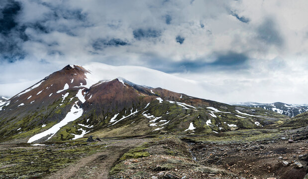 View of Brunavik mountain with snow patches and mossy slopes under a dramatic cloudy sky in Bakkagerdi, Mulathing, Iceland.