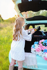 Fototapeta premium Elegant blonde woman serving wine from the open trunk of a compact car decorated with hydrangeas during a warm sunset in nature
