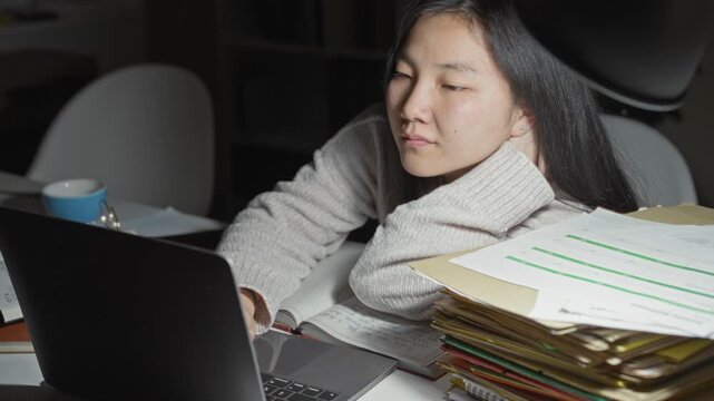 Young woman student rests head on hand while reaching to laptop amid stacked folders, notebook and mug on desk in building; fatigue study concentration.
