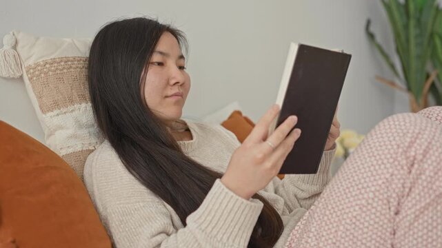 Woman holding book with hand, reclined on bed in building, wearing knit sweater and patterned pajama pants, ring visible on finger while reading and resting; quiet relaxation.