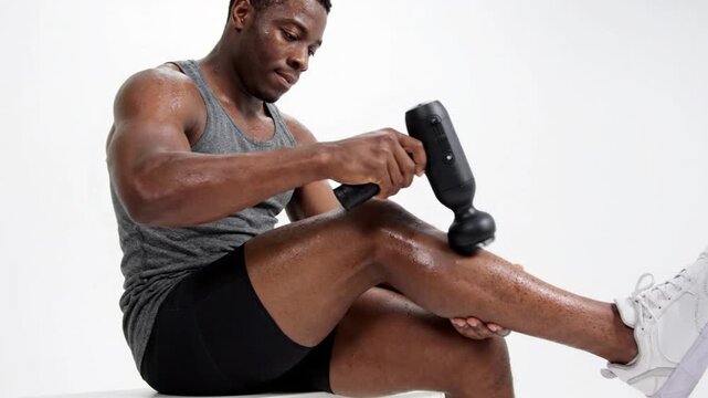A muscular man using a massage gun on his leg after a workout in a modern setting