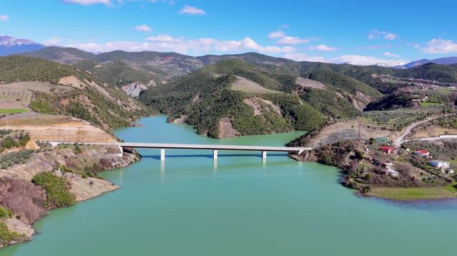 Aerial drone camera moving forward and tilting down over the concrete road bridge crossing the vast turquoise water of Lake Banja reservoir near Gramsh, Albania