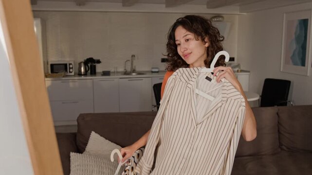 Asian young adult woman comparing two patterned shirts on hangers in front of mirror at home. Footage ideal for fashion vlogging, e-commerce marketing, and morning routine commercials