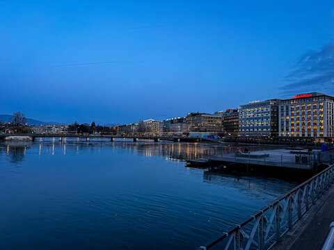 Geneva, Switzerland - 03.09.2026. A tranquil, wide-angle twilight photograph capturing the serene waters of the Rh&ocirc;ne River looking toward Rousseau Island (&Icirc;le Rousseau) and the Mont-Blanc Bridge (Pon