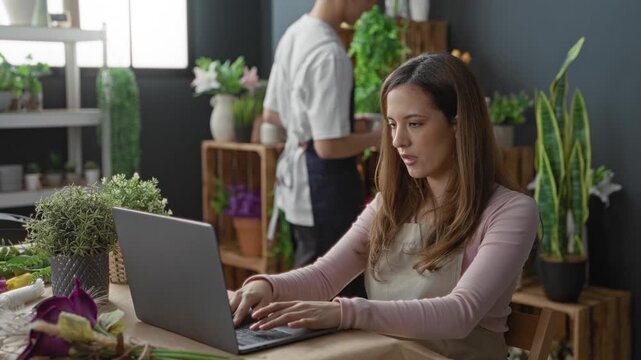 Woman florist typing on laptop, hands on keyboard, wearing apron beside flowers and plants in a studio workspace with man coworker; calm small business.