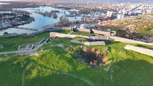 Aerial camera moves forward over the fortified stone ramparts of Rozafa Castle in Shkod&euml;r, Albania, tilting down toward the green grassy terrace during a warm sunset
