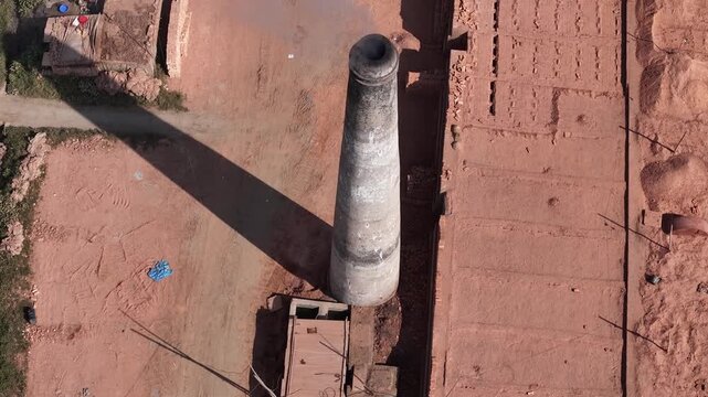 Dhaka, Bangladesh - 13 March 2025: Aerial view of a tall brick kiln chimney casting a long shadow over a brickyard with stacks of red bricks in Dhaka, Dhaka Division, Bangladesh.