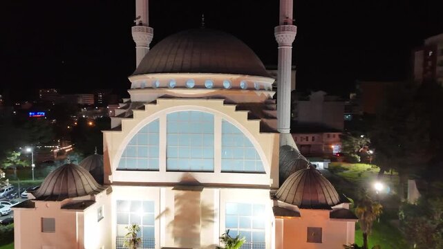 Night drone camera orbiting counterclockwise around the illuminated Ebu Bekr Mosque in Shkod&euml;r, Albania, showcasing its white stepped architecture, main dome, and minaret