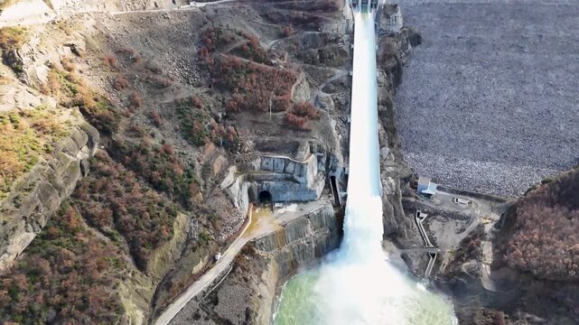 Aerial drone rising while tilting down over the Moglic&euml; Dam spillway in Albania, capturing rushing white water roaring down the concrete channel into the mountain gorge