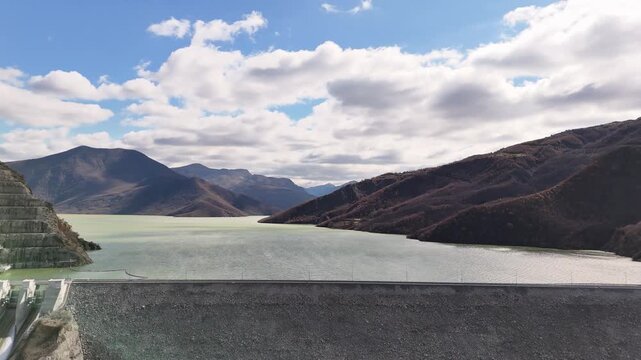 Aerial drone camera flying forward straight over the massive stone Banja Dam wall toward the vast green reservoir lake and rolling mountain valley in Gramsh, Albania