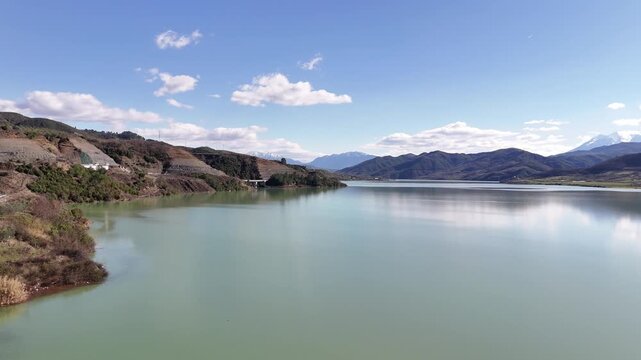 Aerial drone camera tracking left across Lake Banja reservoir in Albania, revealing the pale green water, rugged shoreline slopes, and distant mountains under a blue sky