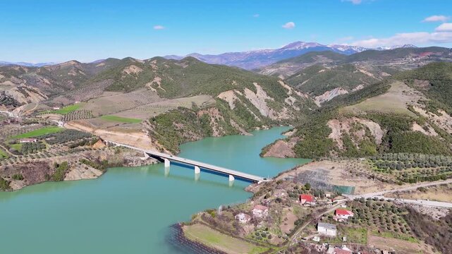 Aerial camera circles clockwise around a modern concrete highway bridge crossing the turquoise waters of Banja Reservoir, surrounded by rolling green hills in Albania