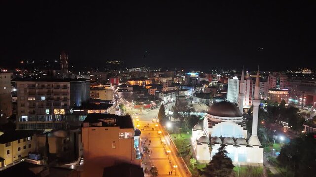 Nighttime aerial drone view over central Shkod&euml;r, Albania, showcasing the illuminated Ebu Bekr Mosque with its two grand minarets, pedestrian promenade, and city skyline