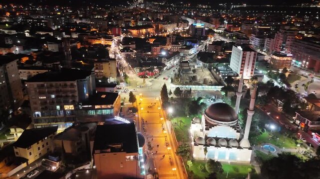 Nighttime aerial drone shot over central Shkod&euml;r, Albania, showcasing the illuminated Ebu Bekr Mosque, its two tall minarets, and the bustling city center city lights