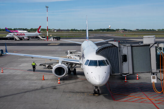 Ground view shows a narrow body jet at a gate with jet bridge at Warsaw Chopin Airport, radar tower near runway, Lauda and Wizz Air jet on stand, crew working at day