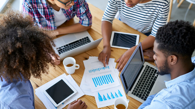 Overhead view of a faceless multiethnic startup team gathered around a wooden table with laptops, tablets and scattered strategy documents, top-down office collaboration concept,