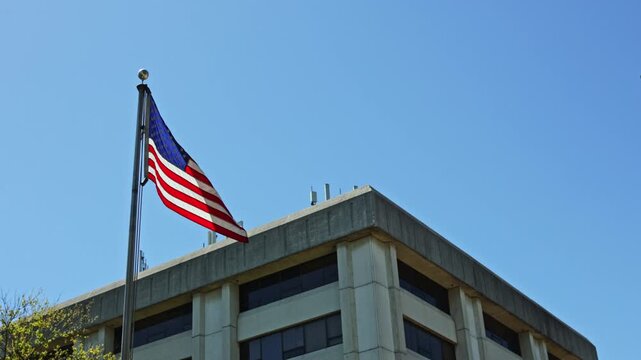 A flag of the United States stands tall above a government office building under a blue sky. The structure features large windows and a flat roof.