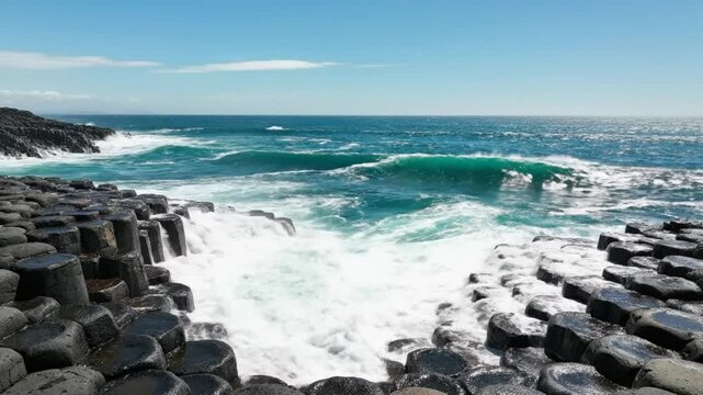 Waves crashing against hexagonal basalt columns with turquoise water under a blue summer sky