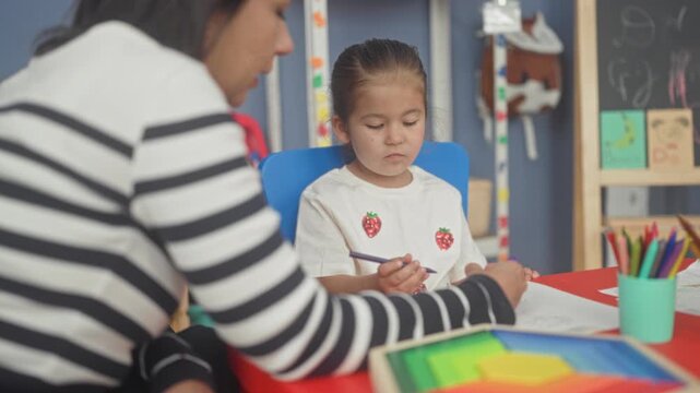 Woman guides girl holding pencil and drawing on paper, points to shapes at red table with colored pencils in classroom building; concentration learning.