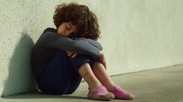 Child curls into ball, head buried, against plain wall. Soft lighting highlights quiet sadness. Could illustrate themes of loneliness, childhood grief, or emotional support needs