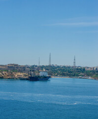 Naklejka premium Cargo ship docked at quay with ancient fortress hillside town and blue Mediterranean sea under clear sky on sunny day in Malta