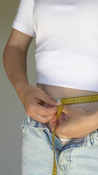 Woman measuring waist circumference with tape measure over loose jeans indoors. Concept of body measurements, slimming progress, fitness challenge and successful healthy diet results.