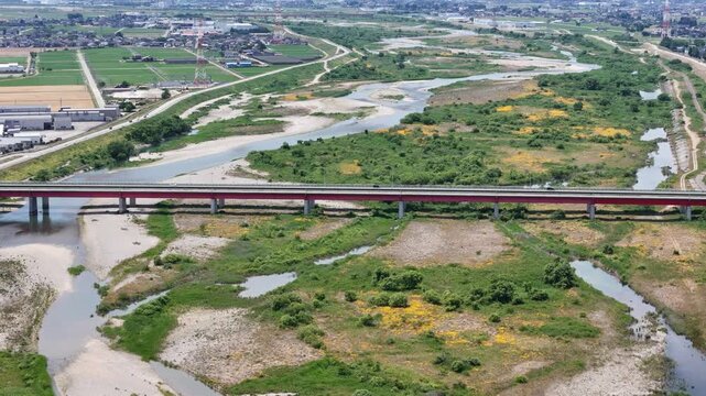 Aerial view of river crossing bridge. Bridge spans wide, winding riverbed. Green patches dot dry, cracked earth. Farmland stretches behind bridge's span. Urban sprawl visible in horizon's distance