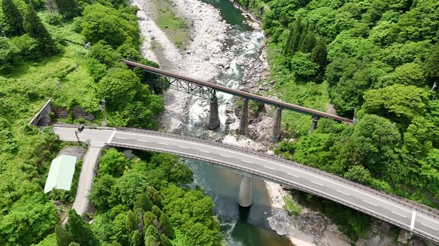 Bridge arches over rushing river, flanked by green forest. Riverbed rocks glisten under sunlight, showing rushing currents. Lone walker moves slowly, admiring the landscape