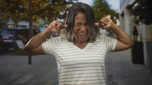 Woman plugs ears with fingers on a city street, eyes closed and face grimacing in discomfort; sensory overload frustration.