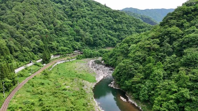 Lush green hills frame a winding river. Train tracks parallel the waterway. Vibrant foliage blankets the mountainside. Nature's serenity invites calm contemplation