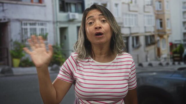 Woman waving hand on a street in front of an apartment building, wearing a striped tshirt with eyes closed and tongue out; dismissive resignation.