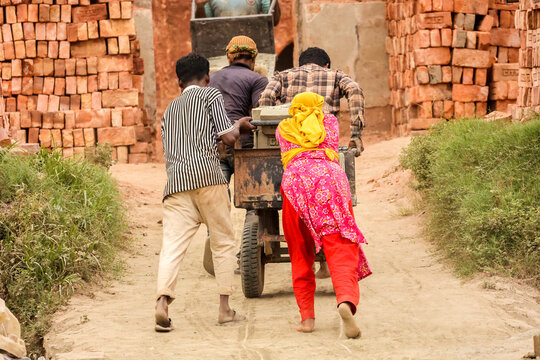 Aminbazar, Bangladesh - 13 March 2026: View of workers pushing a heavy cart loaded with materials through a brick kiln surrounded by stacks of red bricks.