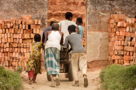 Aminbazar, Bangladesh - 13 March 2026: View of workers and children pushing a heavy cart through a brickyard surrounded by stacks of red bricks.