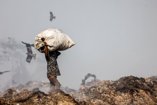 Aminbazar, Bangladesh - 21 February 2026: View of a waste picker carrying a large sack on his head at the Aminbazar landfill with birds flying in the hazy sky.