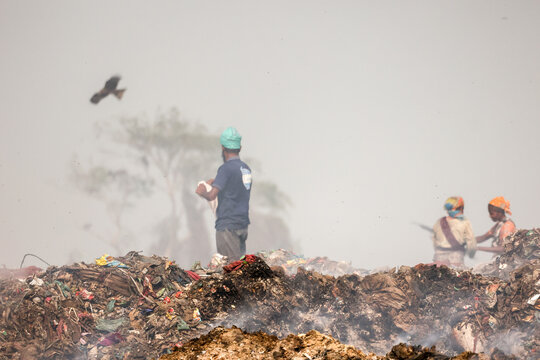 Aminbazar, Bangladesh - 21 February 2026: View of waste pickers working amidst massive piles of garbage at the Aminbazar landfill under a hazy sky with a bird flying.