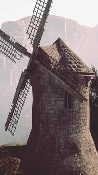 A windmill is in the countryside during the day. The structure has a stone design with large blades. Green land and mountains provide a natural setting, highlighting rural life and history.