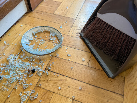 A person sweeps up shattered glass pieces from a parquet floor using a gray brush and dustpan. domestic cleanup and maintenance.