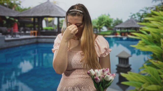 Woman holding tulips pinches bridge of nose near resort pool by building, wearing pink dress with eyes closed and head bowed; quiet sadness.
