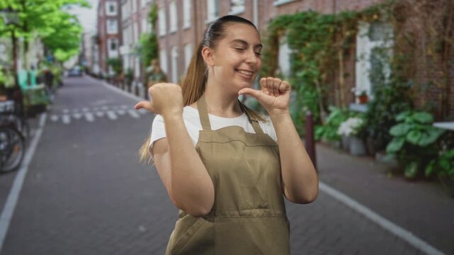 Woman in olive apron pointing thumbs to chest while smiling on a narrow brick street with bicycles and potted plants; pride.