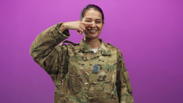 Woman soldier wearing camouflage uniform points finger to nose while showing dogtag and smiling in studio; pride duty.